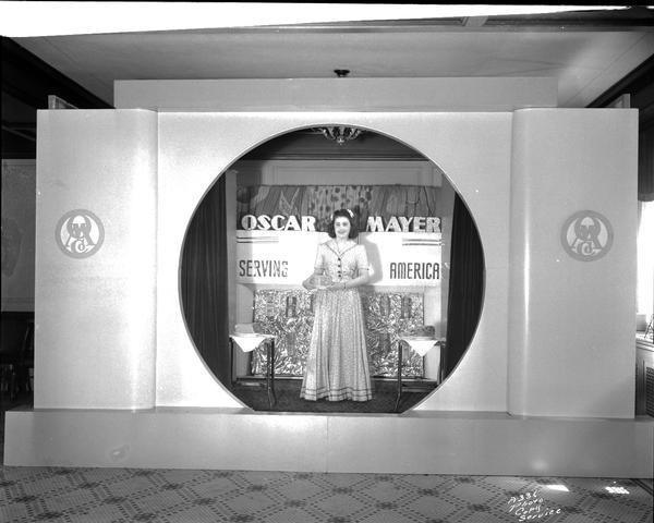 A female model is standing and wearing a long dress on the "Oscar Mayer Serving America" stage set holding a meat product.
