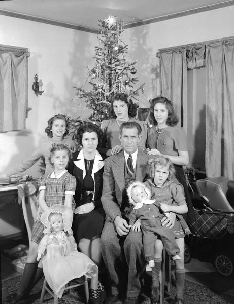 Group portrait of Denton family, showing parents with five daughters, two dolls and a Christmas tree.