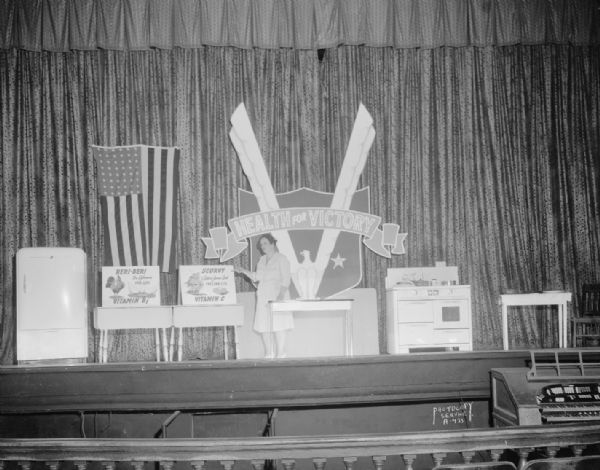 Health for Victory stage, set up with posters, stove and refrigerator for Oscar Mayer at the Eastwood Theater. On the stage is a woman pointing to a poster with a picture of a ship and limes which reads: "Scurvy, Captain James Cook, England-1770 Limes Vitamin C." Another poster with a picture of chickens reads: "Beri-Beri, Dr. Eijkman, Java-1890 Vitamin B1."