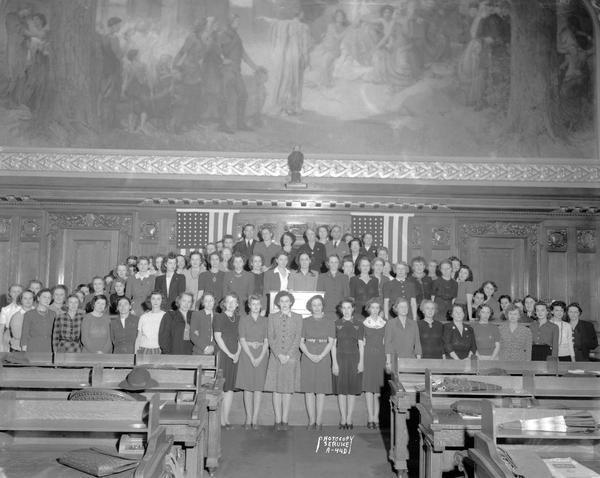 Group portrait of State Civil Service employees enrolled in the staff assistance course for Red Cross and other defense related organizations,  posed beneath mural in the Wisconsin State Capitol Assembly Chamber. Also shows a reproduction of Old Abe, the eagle.