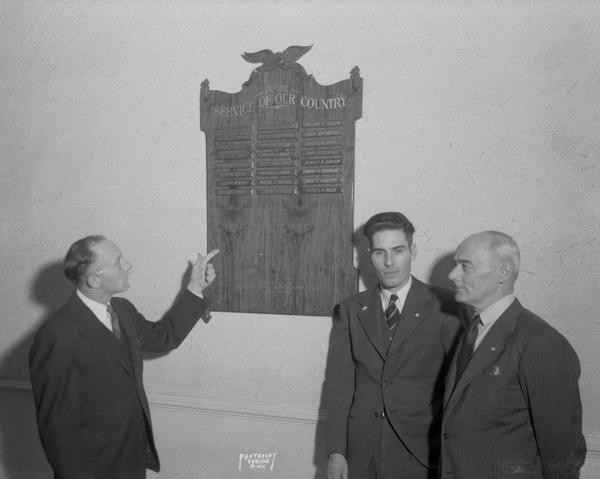 Wooden plaque commemorating 24 Mendota Hospsital employees who are "In the Service of our Country" Plaque was presented by Mendota Hosptial employees Local 13. Standing l to r are: Fred Mundon, Steve Clark, president of Wisconsin State Employees Association and Locxal 13, and Joseph Kozminski, treasurer of Local 13. Jack Welch, head carpenter at the Hospital and member of Local 13, designed the plaque and donated the eagle which was removed from an old style secretary which was used in the old Wisconsin state capitol building.