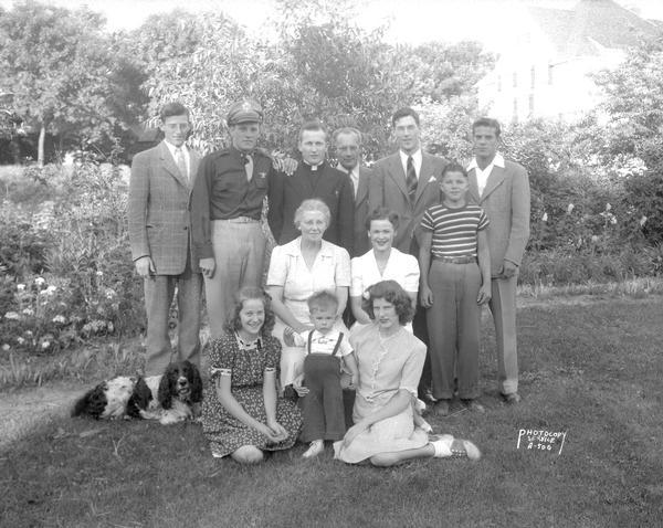 Outdoor group portrait of the John J. Flad Sr. and Nell Flad family. Includes seven adult children, Thomas H., Joseph H., Rev. Fr. Paul N., John J. Jr., James (in uniform), Mary Ellen, and Elizabeth, three grandchildren and a Springer Spaniel dog.