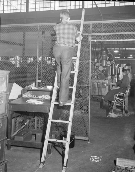 Man standing on folding ladder in Madison-Kipp Corp., with scene of factory floor in the background.