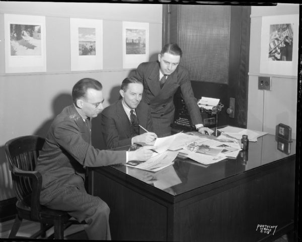Three men are gathered around a desk looking over a copy of <i>The Teller</i>, published by the First National Bank of Wisconsin.