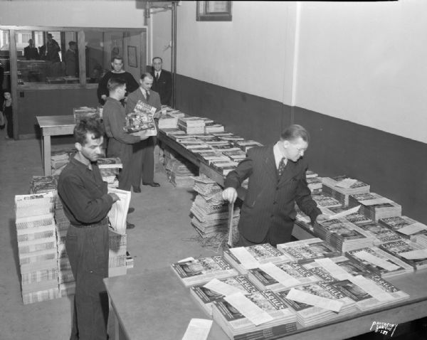 Slightly elevated view of the Madison News Agency, 446 West Gilman Street, work room with six workers sorting periodicals. An office is in the background.