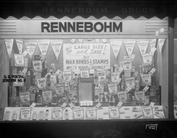 Display of personal care products in the window of Rennebohm Drug Store #10, 676 State Street. Here the promotion to buy large size items seen in other Rennebohm windows in the McVicar-Stein collection is tied to wartime patriotism: "Buy large size and save! Buy War Bonds & Stamps with Your Savings." The fact that a postal sub-station was located in the store made such patriotic purchases convenient to shoppers.