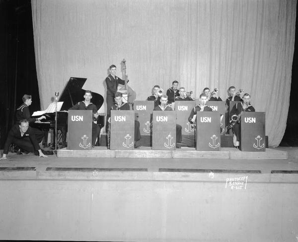 Twelve members of Navy band playing instruments on stage, with a tap dancer posing close to the floor in front of the piano.