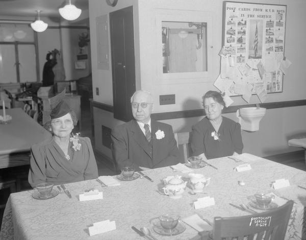 Lena A. Zwank, Theodore J. Schlueter and Mary I. Whare, Wisconsin Motor Vehicle Department retirees, seated at a dining table set up in the office, with postcard display "from MVD boys in the service" in the background.
