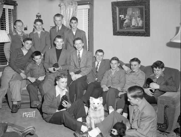Group portrait of 14 young men taken in a living room at a birthday party (for Connie J. Lewis ?), with a dog, and four of the young men holding puppies. Photograph taken for Mrs. Francis Lewis, 1708 Busse Street.