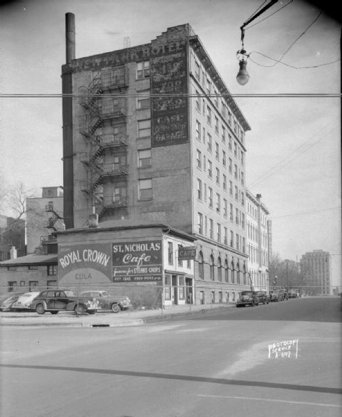 Exterior of Park Hotel, 22 South Carroll Street, rear view, showing St. Nicholas Cafe, 118 West Main Street, with the Tenney Building in the background. Painted on the side of the cafe is a sign for Royal Crown Cola.