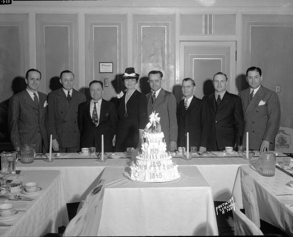 Group of seven men and one woman of the New York Life Insurance Company posing behind a cake celebrating a company milestone at the Pompeiian Room in the Loraine Hotel.