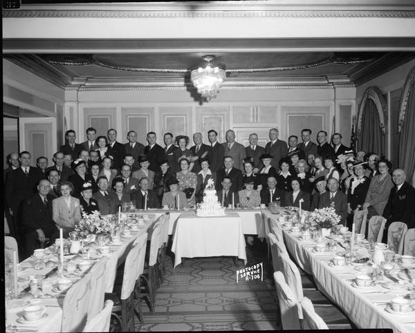 Group of employees of the New York Life Insurance Company posing behind a cake celebrating a company milestone in the Pompeiian Room of the Loraine Hotel.