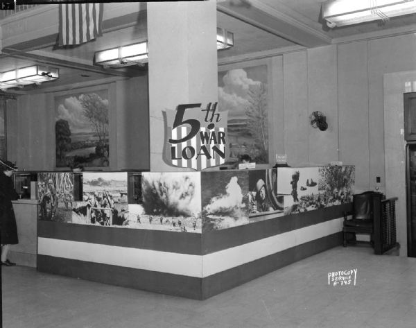 Seven jumbo enlargements of photographs of World War II scenes used to create the War Bond selling exhibit at War Bond Department teller's booths at First National Bank promoting "5th War Loan." Also shown are two large paintings by John Steuart Curry on the wall.