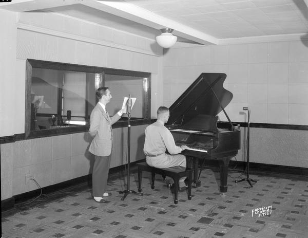 View of the main studio of WIBU Radio Station, 114 North Carroll Street, (Commercial State Bank Building), including one man at the microphone and one man sitting at a grand piano.