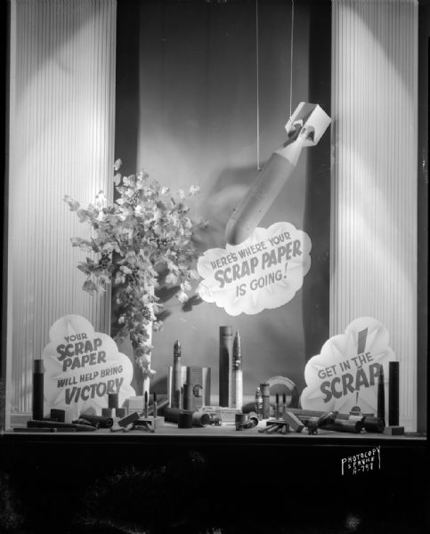H.S. Manchester's, Inc., window display promoting saving paper. Shows fake bomb falling on various ammunition with slogans that read: "Get in the Scrap," "Here's Where Your Scrap Paper is Going!" and "Your Scrap Paper Will Help Bring Victory."