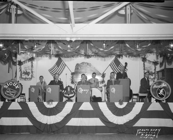 Seven musicians in the Truax Field O. Henry Band on stage, decorated with banners, flags and logos of the Milwaukee Road "Hiawatha" and "Sioux" lines by the Milwaukee Road Employees Service Club, playing for the U.S.O. Carnival Week.