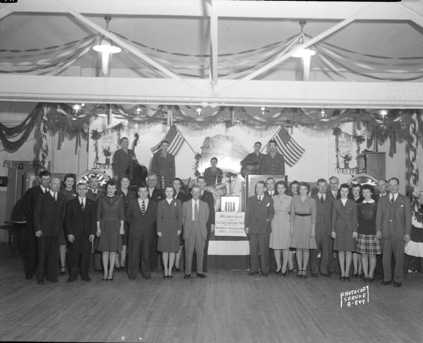 Group portrait of the Milwaukee Road Employees Service Club Committee standing in front of bandstand with musicians. Patriotic decorations provided by the club for U.S.O. Carnival Week.