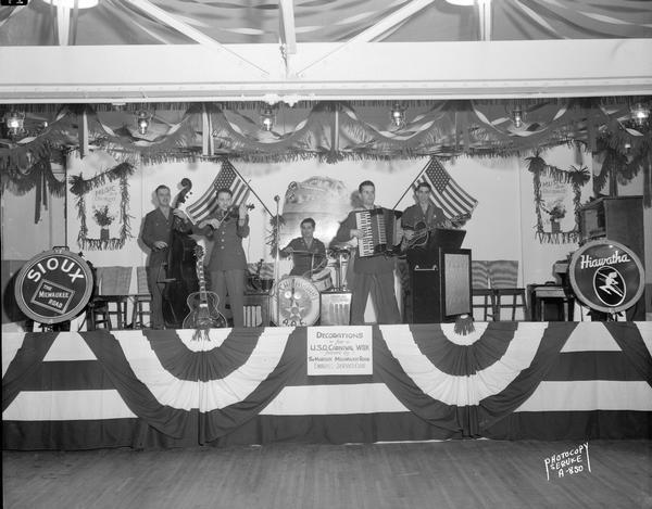 The Army Air Force "Chicagoans" Band, with five musicians playing their instruments on a stage decorated with banners, flags and Milwaukee Road logos for "Hiawatha" and "Sioux" lines for U.S.O. Carnival Week.