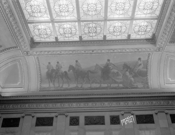 The mural depicts a party of Indians on horseback, on an upland, striking the trail (adapted from a photograph, by permission, made in the National Park). One of four murals by Charles Yardley Turner, representing the evolution of transportation in America. The stained glass skylight in the Wisconsin State Capitol North Hearing Room can also be seen. The title of the mural is: "Indians Striking the Trail."