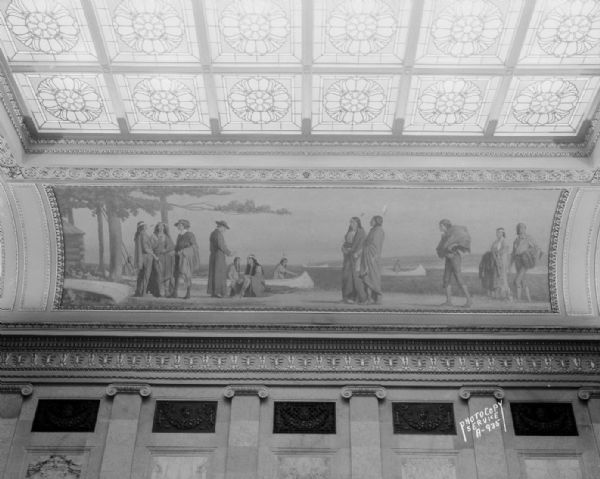 The mural depicts a trading station on the borders of a lake, the Whites and Indians are bargaining for furs, and the canoe is the means of transportation. One of four murals by Charles Yardley Turner, representing the evolution of transportation in America. The stained glass skylight in the Wisconsin State Capitol North Hearing Room can also be seen. The title of the mural is: "A Lake Trading Station."