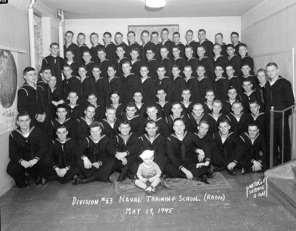 Group portrait of U.S. Naval Training School (Radio), Division #63, with trainees, and a baby boy in sailor suit sitting in front at center, at the University of Wisconsin-Madison.
