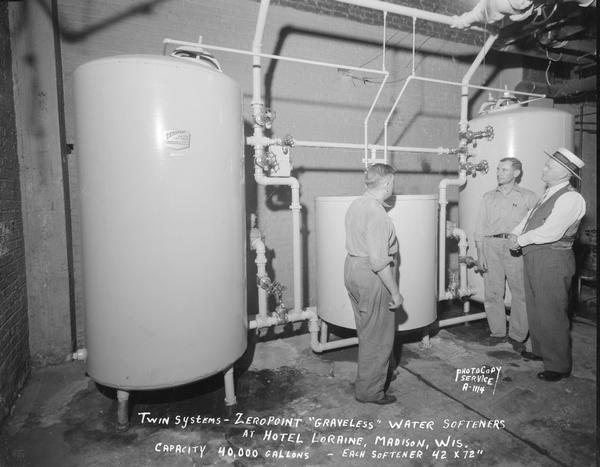 Three men looking at a Zeropoint Water Softener in the basement of the Hotel Loraine, 123 West Washington Avenue. "Twin systems-Zeropoint 'graveless' water softeners. Capacity 40,000 gallons. Each softener is 42 by 72 inches."