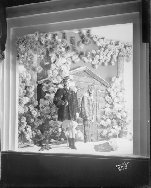 Manchester's, Inc., fur coat window display with two mannequins, one in a fur coat and the other wearing a coonskin cap, standing in the snow in front of a log cabin, taken from Wisconsin Avenue.