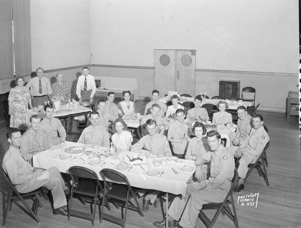 Group of soldiers and young women sitting around two food tables in the Knights of Columbus club room at 15 East Wilson Street.