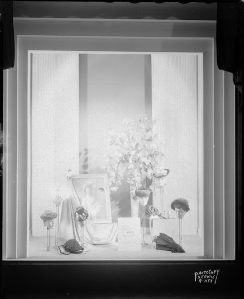 Window #1 at H.S. Manchester's, Inc., 2 East Mifflin Street, showing seven hats, accessories, a floral arrangement, and a portrait of a woman wearing a hat.