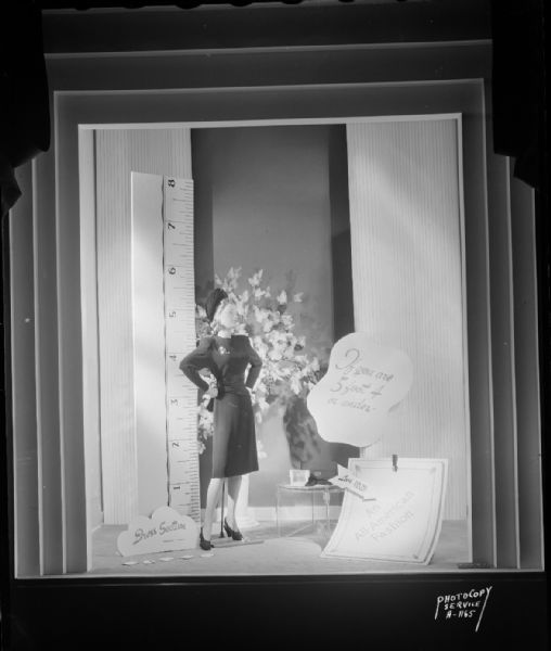 Manchester's, Inc., window #2, "All American Fashion," "If you are 5 feet four or under," "Dress Section, Second Floor." Mannequin in a dress and hat with oversize ruler in the background.