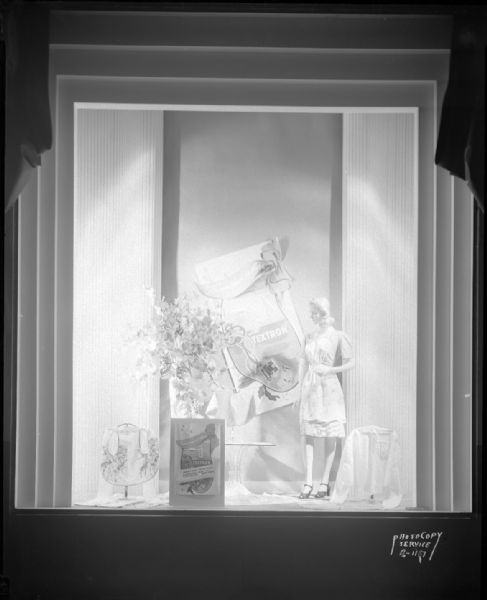 Manchester's, Inc., window #6 featuring "Tomorrow is here with Textron" fabric made into aprons, makeup cape, and blanket bag. The mannequin is wearing an apron.