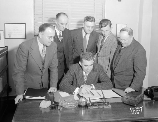 Group portrait of six Madison agents of the New World Life Insurance Company. One man is sitting at the desk, and five men are standing around him, at 110 East Main Street.