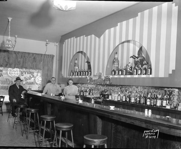 Interior of Glenn-N-Ann's Cozy Inn Tavern, corner of 223 North Frances Street and West Johnson Street, with two bartenders, possibly Glendon and Annadell Hierlmeier, and a customer at the bar. Display of Calvert liquor bottles.
