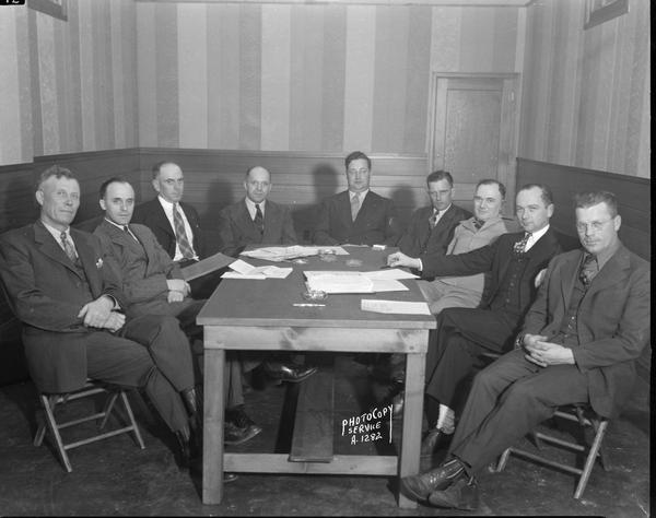 Group of nine men sitting at a table. Taken for Wisconsin R.E.A., 303 East Wilson Street.