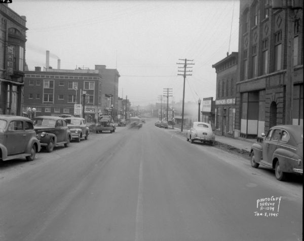 East Wilson Street scene looking northeast, taken at site of Reynolds Bus accident. Businesses shown include R & R Cafe at 316 East Wilson Street, Dizon Men's Clothing at 402 East Wilson Street, Krehl's Drug Store at 408 East Wilson Street, the Cardinal Hotel at 416-18 East Wilson Street, the Milwaukee Road Franklin Street station at 501 East Wilson Street, Howard Machine and Welding Co. at 319 East Wilson Street, and Electric Motor Service at 323 East Wilson Street.