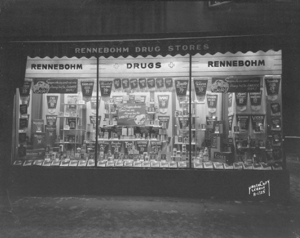 Nighttime view of a window display at Rennebohm Drug Store #7, 901 University Avenue at the corner of Park Street, in the first floor of the First Wisconsin Bank, featuring an array of products for a marketing campaign: "Shoppers who are smart and wise always buy the larger size."