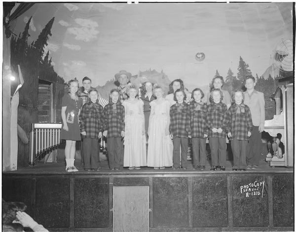 Group of fifteen entertainers, including Don Simon, announcer, three sets of girl twins (Arline and Irene Ripp, Jean and Janette Heinz and Julia and Elizabeth Kalscheuer) in plaid shirts and the Julie and Juliette Gerke twins, on stage at Smitty's Hall, Waunakee, for the grand opening program sponsored by the Milwaukee Cheese Company, which opened a new cheese plant on April 1st.