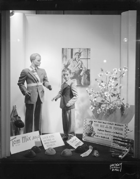 Display window at Karstens Inc., 22-24 North Carroll Street, featuring Tom Mix safety display for the Wisconsin Motor Vehicle Department. The display includes two mannequins, with one wearing a crossing guard bandelier. A picture of cowboy Tom Mix is on the back wall. In the foreground is a series of signs that read: "Tom Mix (of radio) says look both ways before crossing the street," "Don't play in the streets," and "Hey kids! Meet Curley Bradley, Tom Mix, of radio, in person Orpheum Theatre."