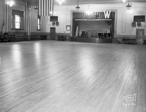 Interior view of the dance hall at the Eagle's Club, 23 West Doty Street. There are chairs and a piano on the stage. Just above the stage is a large "W," and hanging down from the middle of the ceiling is a flag.