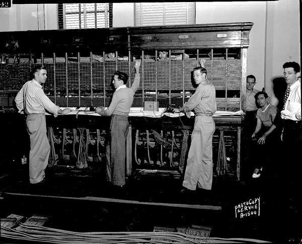 Seven workers (two peeking through from behind the switchboard) and one supervisor rebuilding the telephone toll switchboard at the Wisconsin Telephone Company building.