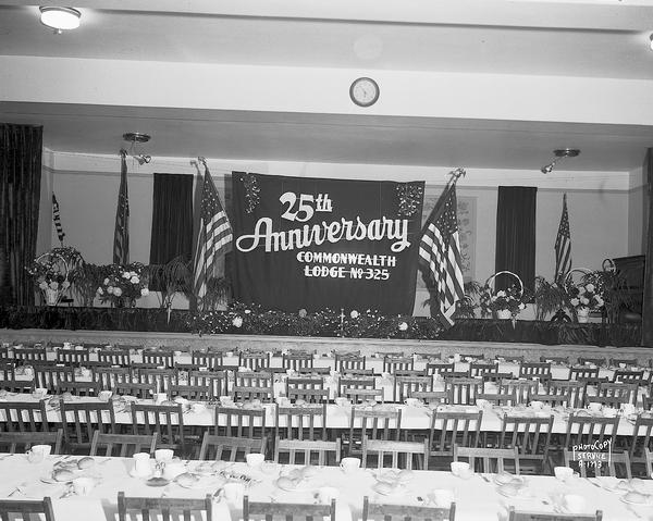 Masonic Commonwealth Lodge No. 325 twenty-fifth anniversary dinner, showing empty dining hall, floral decorations on speakers table, and American flags behind the speakers table.
