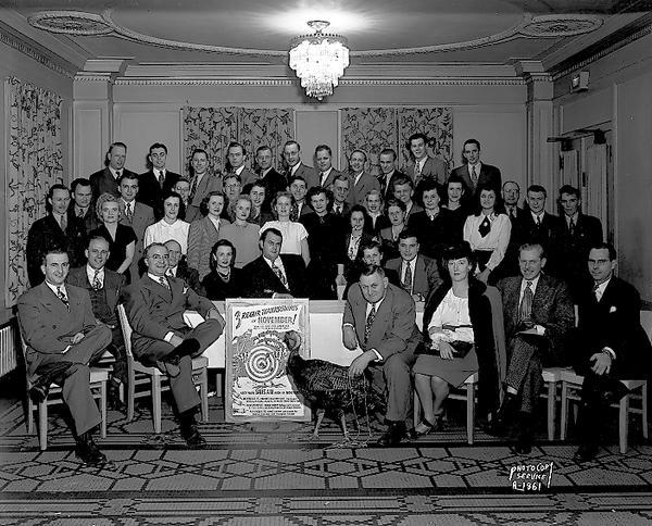 Group portrait of Rexair (vacuum cleaner) Company male and female employees in the Pompeii Room of the Loraine Hotel, with a live turkey and a poster that says: "Three Rexair Thanksgivings in November, keep your sales high in November."