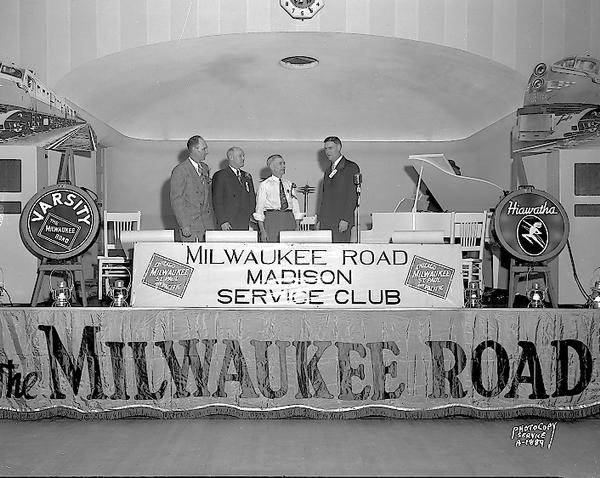 Milwaukee Road Service Club, with four men on stage with a banner along the front that reads: "Milwaukee Road Service Club." VFW Hall, 113-115 E. Mifflin Street.