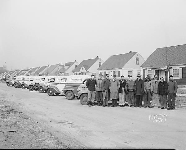 Line-up of six Pettersen's Inc. delivery trucks. Sign on trucks: "Rugs, Carpets, Linoleum." Also shows 2 cars and 11 employees. Includes a view of the 2600 block of Myrtle Street.