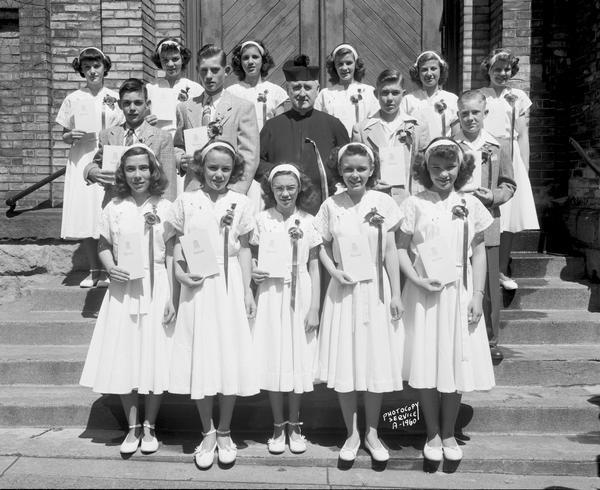 Group portrait of Saint Patrick's School graduation class taken on the front steps of Saint Patrick's church at 410 East Main Street. Includes eleven girls, four boys, and one priest.