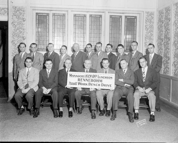 Group portrait of eighteen Rennebohm Drug Store managers holding a sign "Managers' Kick-Off Luncheon, Rennebohm, Rexall Work Bench Drive."