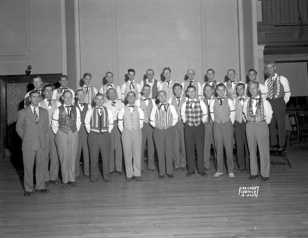 Group portrait of twenty-six SPEBSQSA (Society for the Preservation and Enjoyment of Barber Shop Quartet Singing in America) members wearing colorful vests, and their director, who is wearing a suit.