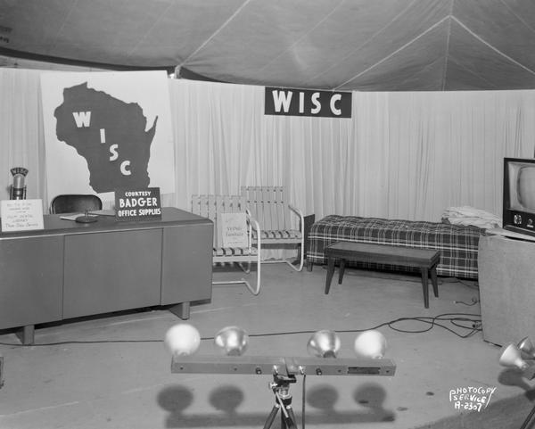 WISC television studio set-up at Parade of Home, featuring desk and informal seating and sign that reads: "All TV film shown here is from the film rental library, Photo Copy Service." 
