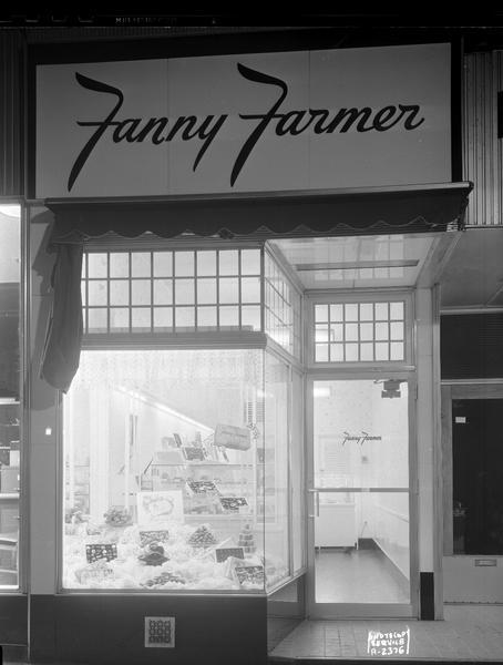 Exterior view at night of Fanny Farmer Candy Store, 10 South Carroll Street, including a display window featuring a variety of candy.