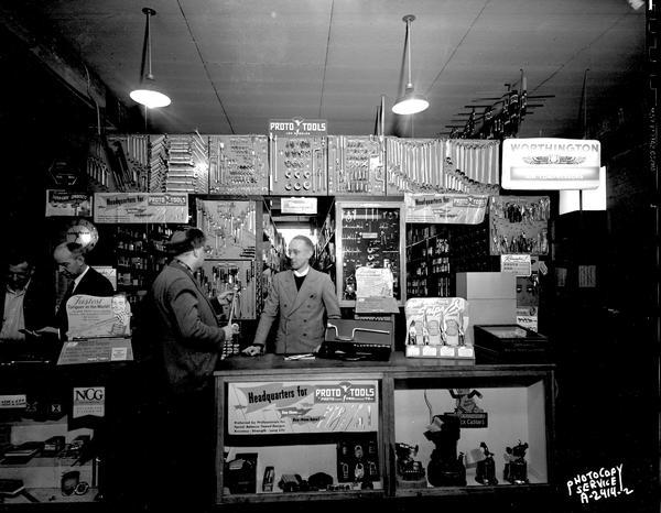 Interior of Richard Ela Company, 744 Williamson Street, showing a sales counter with a large display of tools and three clerks and one customer. Alternate view.